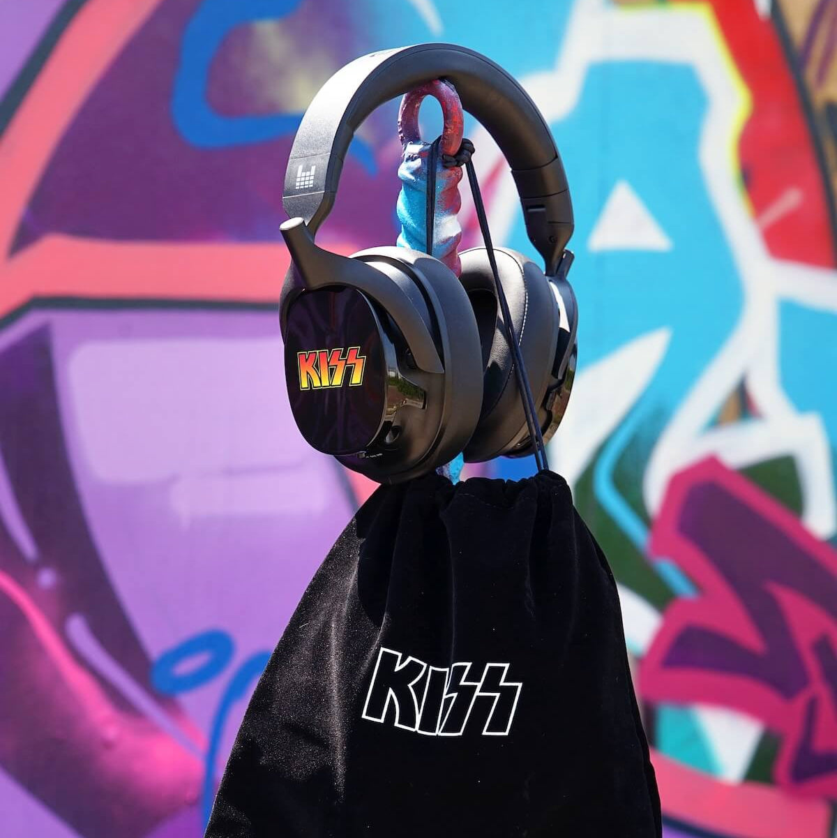 Black over-ear headphones with a KISS logo on the earcup, hanging from a pink rope above a black drawstring pouch with KISS branding, set against a colorful graffiti wall.